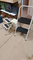 Two metal two-step stools, plastic step stool, and plastic garbage can grouped together on basement floor.