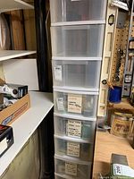 Nine clear plastic storage drawers stacked one on top of the other next to a workbench and shelving in basement area.