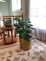 Living room scene showing the large faux plant with broad green leaves inside a brass-colored metal planter on a floral carpet near a window and lace curtain.