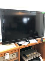 Front view of Sony Bravia TV on wooden desk with remote and manual visible below screen.