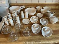 Wide view showing all items in the kitchenware lot on wooden countertop, including ceramic dishes, egg holders, creamers, salt and pepper shakers, and glass cloches.