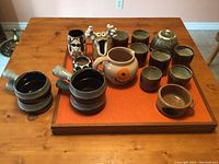 Overview of all ceramic items on orange tray atop wooden table showing mugs, cups, pitcher, and decorative pieces