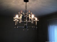 Metal and crystal chandelier lit with six candle-style bulbs in a room with neutral walls and window with sheer curtain.