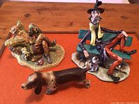 Photo showing all three figurines together on wooden table: reclining man with pipe, seated man on green bench with scarf and dog, and separate dog figurine in front.