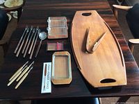 Image shows a Fondue Plus incomplete set with wooden serving tray, fondue forks, glass dishes, metal spoon, wooden tongs, and a box of After Fondue forks laid out on a dark wood table.