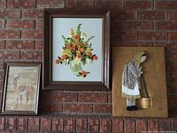 Three framed art pieces hanging on a brick wall; needlepoint woman, floral bouquet, and deer print.