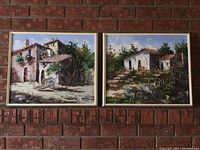 Two framed art prints hanging on a brick wall showing Mediterranean village houses with detailed brush strokes and signed 'Pepe'.