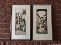 Two framed watercolor paintings depicting urban street scenes, hung on a brick wall.
