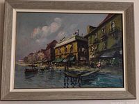 Framed painting showing a scenic coastal village with boats and buildings by the water under a partly cloudy sky.