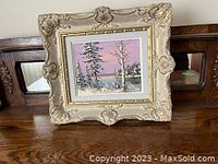 Full view of the framed artwork placed on a wooden surface, showing the ornate cream-colored frame and winter landscape painting inside.