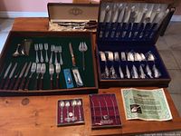 Photo showing various flatware and cutlery pieces arranged in trays and cases, including boxed sets and loose pieces.
