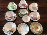 Nine assorted vintage porcelain teacups with matching saucers laid out on wooden surface, various floral designs and colors.