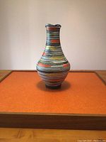 Front view of ceramic vase showing multicolored horizontal stripes and orange rectangular accents on an orange background surface.