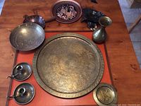 Photo showing 7 brass and metal decorative items laid out on wooden table including large engraved tray, bowls, small plates, candle holders, and a vase
