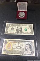 Silver proof dollar coin in red velvet-lined case along with US and Canadian one dollar bills placed on black surface.