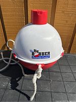 Front and top view showing logo 'The Big Bobber Floating Cooler', red bottom half and white top half, with attached white rope.