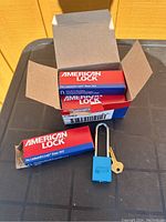 Open box showing multiple sealed boxes of American Lock padlocks, one blue padlock with extended shackle and one key outside.
