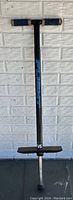 Full view of Flight pogo stick standing against a white brick wall, showing blue grips, black shaft, and foot pegs.