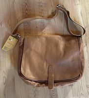 Tan leather messenger bag laid flat on wooden floor showing front flap and adjustable shoulder strap with original tag attached.