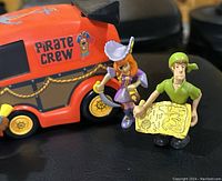 Close up photo showing Scooby Doo figure, female pirate figure, male pirate figure with treasure map, and the side of the pirate crew van with red-orange color and graphics.