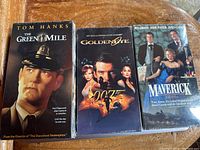 Front view of three sealed VHS tapes The Green Mile, GoldenEye, and Maverick arranged side by side on a wood surface.