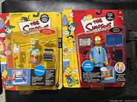 Photo showing two sealed Simpsons action figures: Wendell and Herb Powell, in original packaging with accessories and colorful background art.
