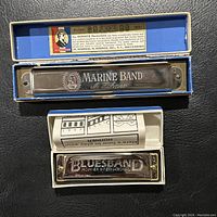Top view of two harmonicas side by side; vintage Marine Band in blue box and newer Bluesband model in white box, showing branding and original packaging.