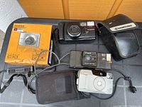 Four cameras arranged on a flat surface including a boxed Fujifilm FinePix C10, Samsung camera with case, Canon Sure Shot Supreme, and Canon Prima Zoom 80 with case.