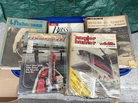 Photo showing an assortment of vintage fishing magazines with visible titles including The Angler and Hunter, Rod and Gun in Canada, and American Angler mostly in plastic preserving bags on top of a blue container.