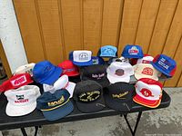 Wide view of 19 vintage fishing trucker hats on black table against wood wall, showing various colors and logos related to fishing.