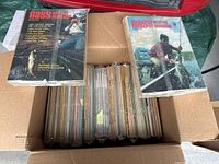 Open cardboard box filled with vintage Bass Master Magazines from 1982 to 2000 with two magazines placed on top showing varied cover designs and formulas.