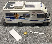 Side view of the white Majorette Holiday Camper motorhome toy with three small detached pieces placed beside it, showing the design and condition.