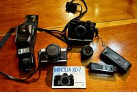 Two vintage Minolta cameras and accessories arranged on wooden surface including a Minolta XD-7 SLR camera, a Minolta compact camera, flashes, lenses, camera straps and owner's manual.