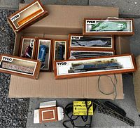 Overview of the lot showing multiple boxed Tyco train cars, curved track sections, and Lionel transformer on cardboard.