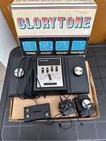 Top view of Glorytone Video Olympiad console with two paddles, wired accessories, and original box in background labeled Tennis, Hockey, Squash, Practice.