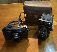 Two black vintage Yashica cameras on wood surface with brown leather carrying case, Yashica MG-1 and Yashica-24 TLR.