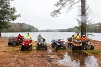 Five ATVs parked lakeside with riders wearing helmets and outerwear