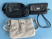 Top-down view of three handbags arranged on blue fabric: black Halston bag, black Nine West bag with chain strap, white Aldo bag with buckle front.