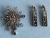 Photo showing front view of Guy Vidal modernist brutalist brooch and pairs of dangle earrings on a blue background.