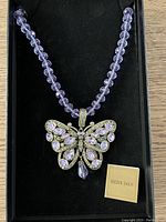 Full view of purple glass bead necklace with butterfly pendant inside a black box with Heidi Daus branding and product information card.