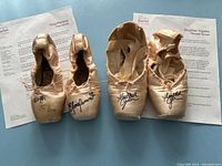 Two pairs of ballet pointe shoes placed side by side on a white surface with information sheets behind each pair. One pair on the left signed by Tina Pereira and the other pair on the right signed by Heather Ogden.