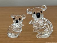 Front view of two Swarovski crystal koala figurines on a wooden surface, showing clear faceted crystal and black nose/eyes.