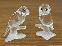 Front view of Swarovski crystal owl and parrot figurines on wooden surface showing clear faceted crystal with frosted base and colored eyes.