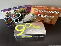 Photo showing all three Trivial Pursuit editions: 80s Totally (purple box), 90s Time Capsule (metallic tin), Pop Culture 2 DVD (brown box)