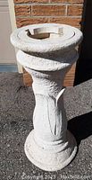 Photo of the white textured resin bird bath base standing outdoors against a brick wall, showing the front side with the circular top and decorative vertical leaf design