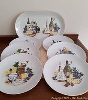 Seven porcelain items: six round appetizer plates and one oval platter with retro styled grape, cheese, and wine bottle illustrations. Plates stacked and platter behind.