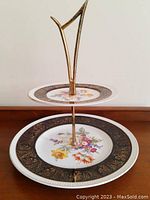 Full view of vintage two tier cake stand showing bottom and top plates with floral patterns and black gold border, supported by brass handle