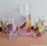 Front view of the glass pitcher and four juice glasses with colorful fruit patterns on a table surface.