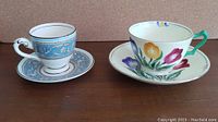 Two vintage Staffordshire tea cup and saucer sets shown side by side on a wood surface: one set with blue floral demi tasse cup and saucer, the other with white cup and saucer decorated with tulip flowers in various colors.