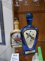 Two Beam whiskey bottles displayed on a glass shelf, one blue with duck artwork and one cream with a cowboy painting.